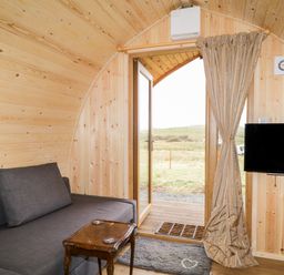 A small wooden pod interior with a gray couch a small wooden side table a wall-mounted television and a door opening to a grassy outdoor area at Garraidh Ghorm Pod in Conista