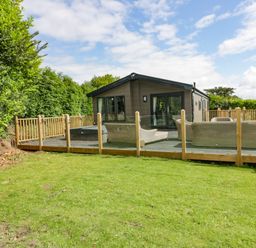 A wooden house with glass doors and outdoor seating on a fenced deck surrounded by grass and trees at Pinehurst Lodge in St Day