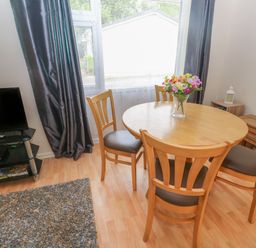 A dining area with a round wooden table and four chairs with a vase of flowers on the table next to a window with dark curtains a television on a stand and a rug on the floor at 11 Tremore in Liskeard