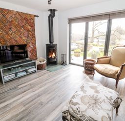 A living room with a TV on a stand a patterned wall hanging a wood stove an armchair and glass doors showing an outdoor view at Romsey Lodge in Wark
