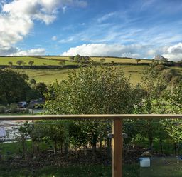 A view of green fields and trees seen from behind a wooden balcony railing at 1 Valley View in Lanreath