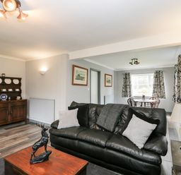 A living room with a black leather sofa and cushions a wooden coffee table with a deer statue a side table with a lamp and sculpture a wooden cabinet with china plates and cups a dining table with chairs near windows with curtains at Pool House in The Narth near Trellech