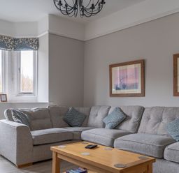 A living room with a grey sectional sofa blue patterned cushions wooden coffee table two framed pictures and a window with patterned valance at Keepers Cottage in Hawkshead