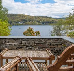 A stone patio with wooden chairs and a table overlooking a lake and wooded hills at Grassholme in Hawkshead