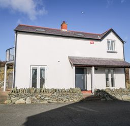 An exterior view of a two-story house with white walls and a stone fence at Bay View in Trearddur Bay