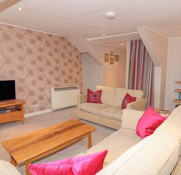 A living room with beige sofas and pink cushions a wooden coffee table a tv on a wooden stand floral wallpaper and bookshelves at Manor Studio in Dartmouth