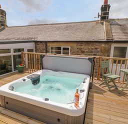 A hot tub on a wooden deck outside a stone house with a table and chairs at Crag View in West Woodburn