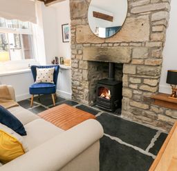 A living room with a stone fireplace and wood stove a beige sofa with pillows a blue chair and a wooden table at Pine Cottage in Sedbergh