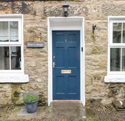 A stone exterior wall with a blue door numbered 1 a plant pot and two windows at Pine Cottage in Sedbergh