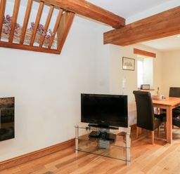 A dining area with a wooden table and four black chairs next to a glass TV stand with a television at Chapel Cottage in Leonard Stanley near Kings Stanley