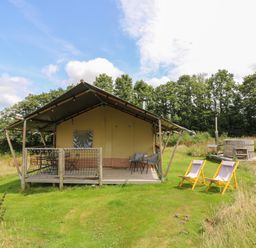 A glamping tent with a wooden deck, outdoor chairs, and a hot tub in a grassy area with trees at Honey @ Stars & Embers Glamping in Grampound