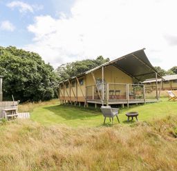 A raised tent structure with a covered porch and outdoor furniture on a grassy area near wooden hot tub and fire pit at Bumble @ Stars & Embers Glamping in Grampound