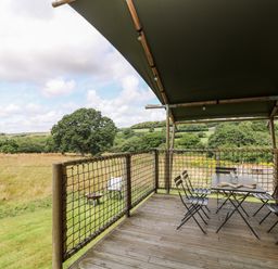 A wooden deck with a table and chairs overlooking a grassy field and trees at Bumble @ Stars & Embers Glamping in Grampound