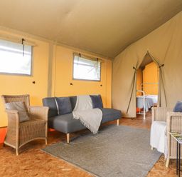 An interior of a tent with a sofa, two wicker chairs, a small table with cups and magazines, and a view of a bedroom with a bed at Bumble @ Stars & Embers Glamping in Grampound
