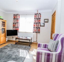 A living room with a striped purple armchair a wooden cabinet a mounted television and a coffee table near a window with patterned curtains at Lillies Pad in Methwold near Feltwell