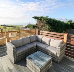 An outdoor seating area with a gray cushioned sectional sofa and a matching glass-top coffee table on a wooden deck at Caban Tywod in Abersoch
