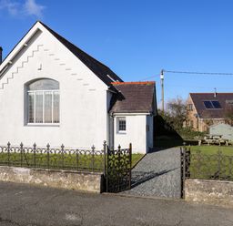 A white building with a black roof and metal fence with a garden bench and shed behind it at Capel Hermon in Llanynghenedl near Bodedern