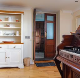 A room with a wooden floor a white cabinet with pottery and mugs an open wooden door and a wooden piano at Capel Hermon in Llanynghenedl near Bodedern