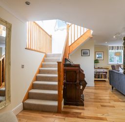 A carpeted staircase with wooden railing next to a vintage wooden desk in a hallway opening into a living room with bookshelves and a gray sofa at Capel Hermon in Llanynghenedl near Bodedern
