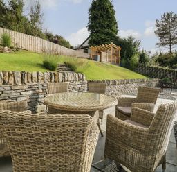An outdoor seating area with six wicker chairs around a glass table on a patio next to a stone wall and grassy slope at Bishop Fell in Newby Bridge
