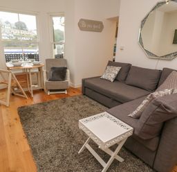 A living room with a gray sofa, a white tray table, a round dining table with chairs near windows at 1 River View in Looe