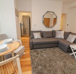 A living room with a gray corner sofa a round wooden dining table with place settings and a refrigerator visible in the doorway at 1 River View in Looe