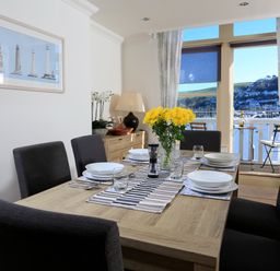 A dining room with a wooden table set for six with flowers on the table and a view of water and hills through windows at 2 Embankment House in Dartmouth