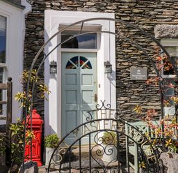 The entrance door of a stone house with a metal arch and gate with the words The Masters House a red mailbox and plants at The Postmasters House Windermere