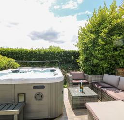 An outdoor patio with a hot tub a wicker sofa set and a glass table with a bottle and glasses at Trevore Farmhouse in Chacewater