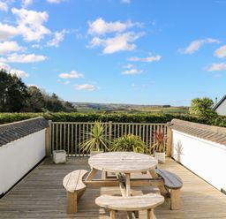 An outdoor wooden deck with a round picnic table and benches with potted plants and countryside views at Trevore Farmhouse in Chacewater