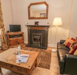 A living room with a black sofa with cushions a wooden coffee table with magazines a fireplace a wooden TV stand with a TV and a floor lamp at Manod View in Blaenau Ffestiniog