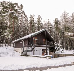 A cabin in a snowy wooded area with outdoor chairs and a pathway covered in snow at Torbreck Chalet in Kincraig