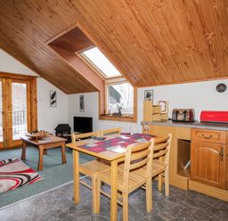 A dining area with a wooden table and chairs next to a kitchen countertop and a living area with a coffee table chess set and a television at Torbreck Chalet in Kincraig