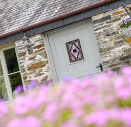 A stone cottage exterior with a grey door featuring stained glass and a window with curtains near a hanging flower basket at Mimi's Cottage in St Neot