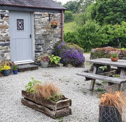 An outdoor patio area with a wooden picnic table and benches surrounded by potted plants and a stone building with a painted door at Mimi's Cottage in St Neot