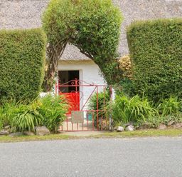 A red metal gate with an archway of trimmed greenery leading to a white building with a thatched roof at Mary Rose Cottage in Cordal near Castleisland County Kerry