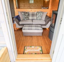 An open doorway leading to a wooden cabin interior with a sofa, matching ottoman, wood stove, and welcome mat at Badgers Sett in Winston near Staindrop