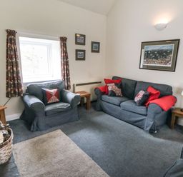 A living room with a blue armchair and sofa with red and patterned cushions a wooden side table a TV and a basket of firewood at Silver Birches in Glaisdale