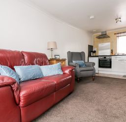 A living room with a red leather sofa blue cushions a grey armchair a wooden side table with a lamp and a kitchen area at Coach House Cottage in Bude