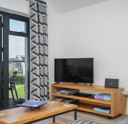 A living room with a wooden coffee table and TV unit next to a glass door with patterned curtains at Devonair in Westward Ho