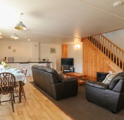 A living and dining area with a wooden dining table set with dishes and cutlery leather sofas a coffee table a television under stairs and kitchen appliances at The Dairy in Bude