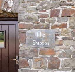 A stone wall with a sign that says The Shippon next to a brown wooden door at The Shippon in Bude