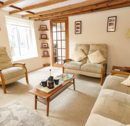 A living room with beige armchairs a wooden coffee table with a tea set and magazines a standing lamp and a window at Primrose Cottage in Stiffkey