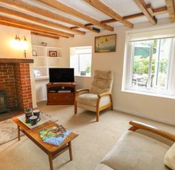A living room with wooden beams on the ceiling a brick fireplace a television on a wooden stand an armchair a sofa a wooden coffee table with cups and magazines and windows showing a garden at Primrose Cottage in Stiffkey