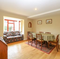 Dining room with a table covered by a green polka dot tablecloth surrounded by six wooden chairs and a floral patterned sofa by a window at Primrose Cottage in Stiffkey