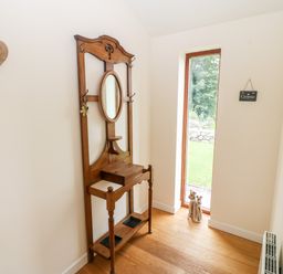 A hallway with a wooden hall tree featuring an oval mirror a small shelf and hooks next to a tall narrow window with a garden view at Llofft Yr Yd in Llandwrog near Dinas Dinlle
