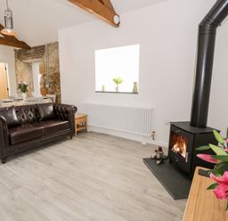 A living room with a brown leather sofa wood burning stove and flowers on a table at 1 Mountain View in Talwrn near Llangefni