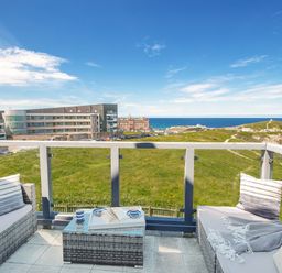 A balcony with wicker furniture and cushions overlooking a grassy area buildings and the sea at Breakers in Newquay
