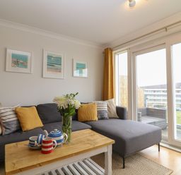 A living room with a gray sectional sofa yellow and striped cushions a wooden coffee table with a tea set and flowers near large windows at Breakers in Newquay