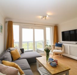 A living room with a grey sofa and yellow curtains overlooking a balcony at Breakers in Newquay